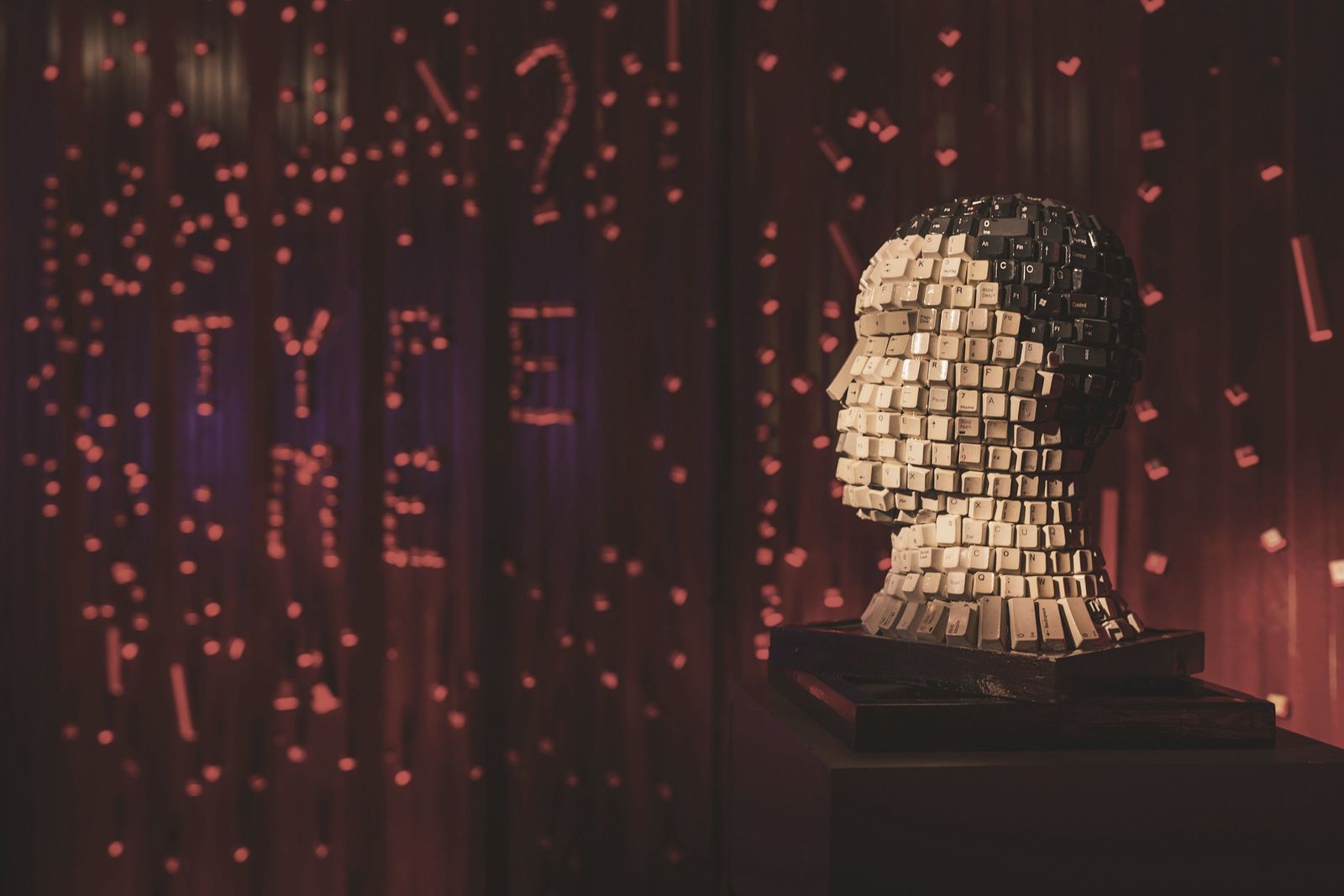 A sculpted head made of keyboard keys, lit red, with TYPE ME spelled out in lights behind it