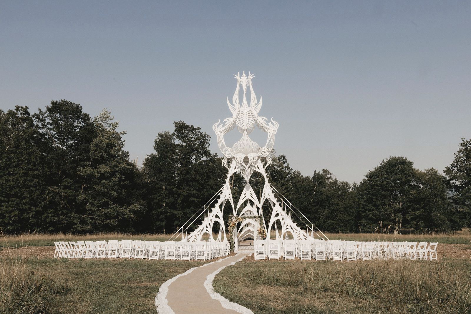 A sculptural lace wedding altar at the end of a winding aisle, with rows of empty white chairs in a meadow