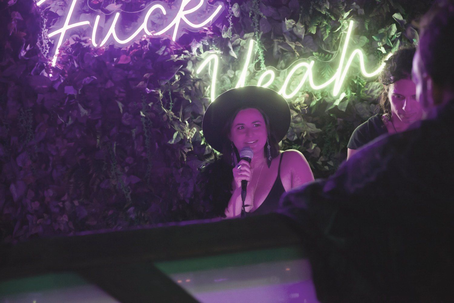Jade on the mic in a wide-brimmed hat beneath a neon sign reading Fuck Yeah