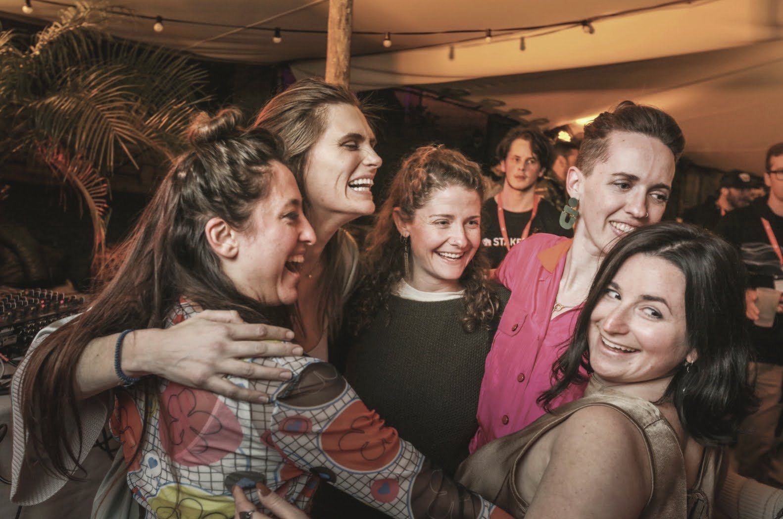 Five friends in a laughing group hug under string lights