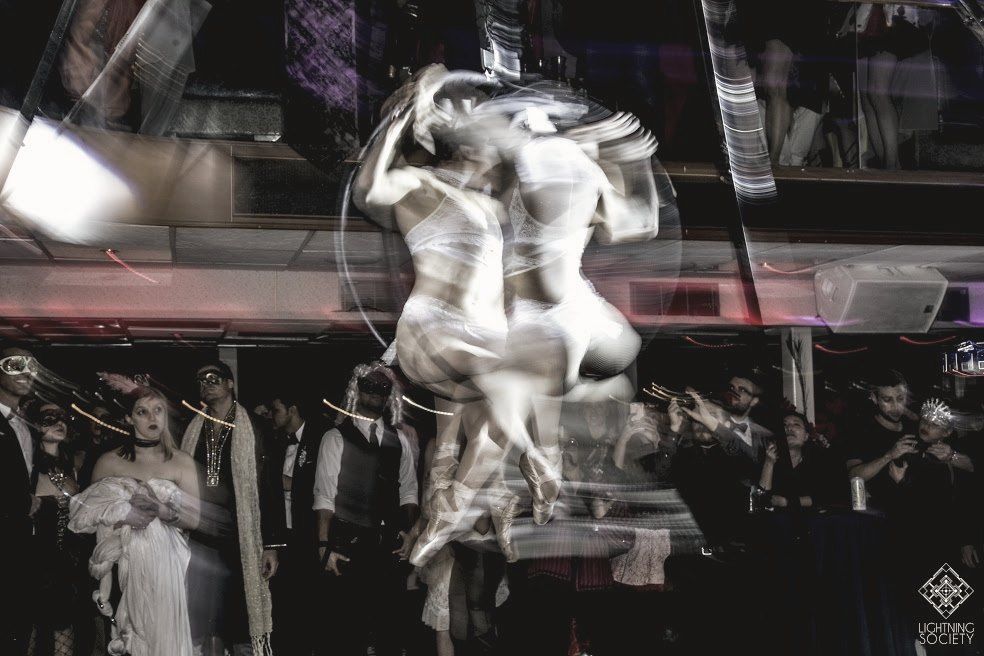 An aerialist spinning mid-air above a masked, costumed crowd, blurred with motion
